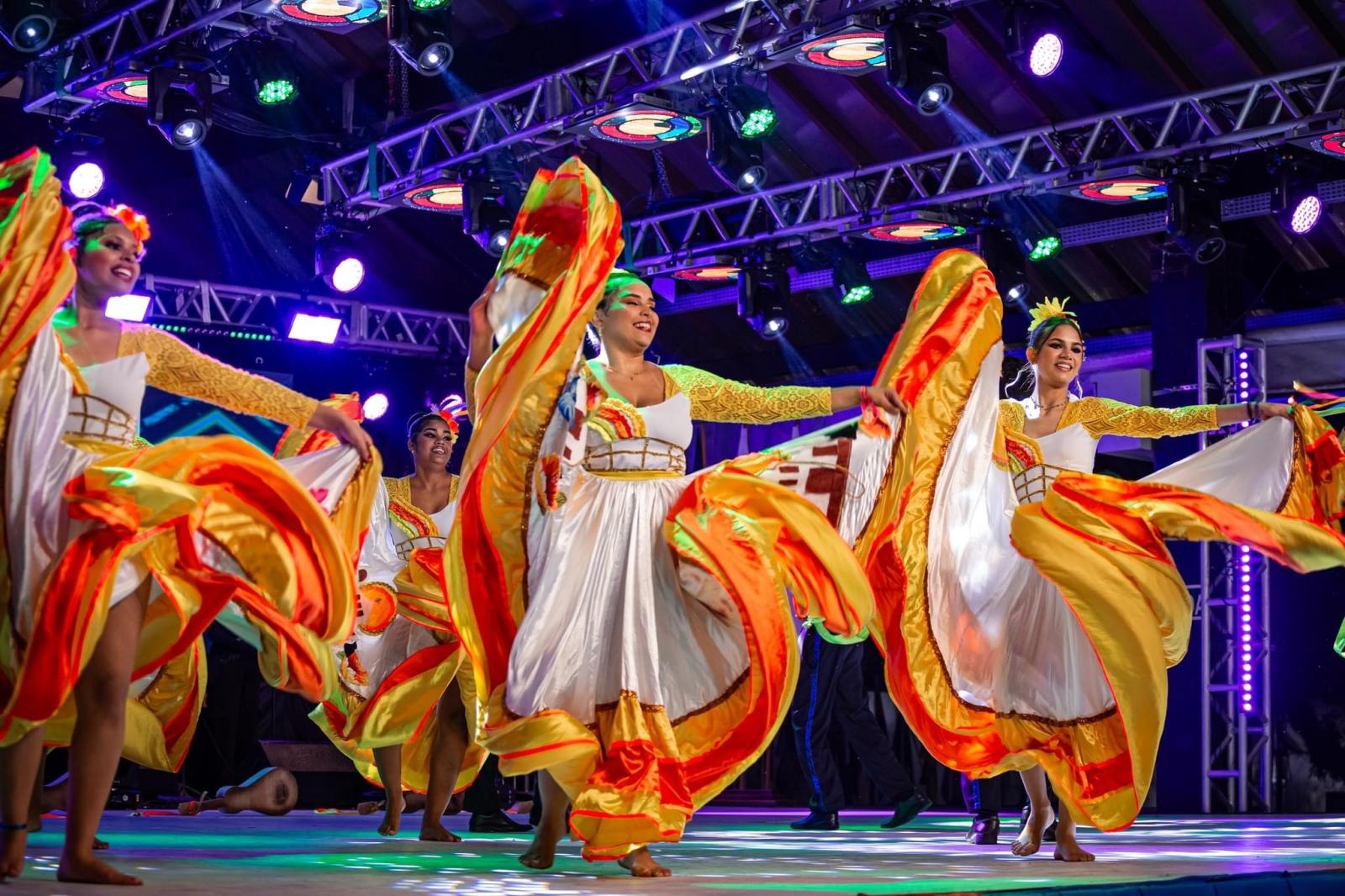 Grupo de dança Flor de Atalaia, do Mato Grosso do Sul. A foto mostra quatro moças em um palco, dançando com vestidos brancos de saia rodada com detalhes em amarelo e vermelho.