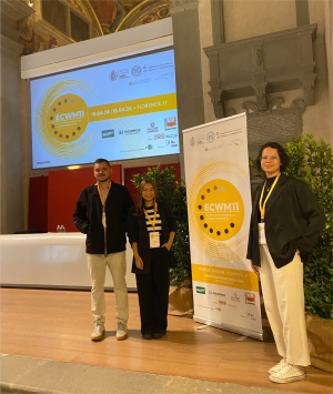 Foto dos estudantes Saulo, Kamilly e Anna Clara (da esquerda para a direita), em pé ao lado de um banner do evento.