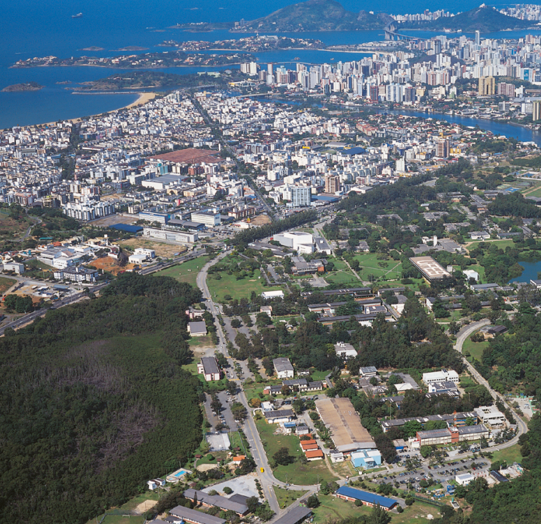 Fotografia aérea do campus de Goiabeiras, com suas áreas verdes e prédios, tendo ao fundo parte da cidade de Vitória e o mar em tom azul escuro