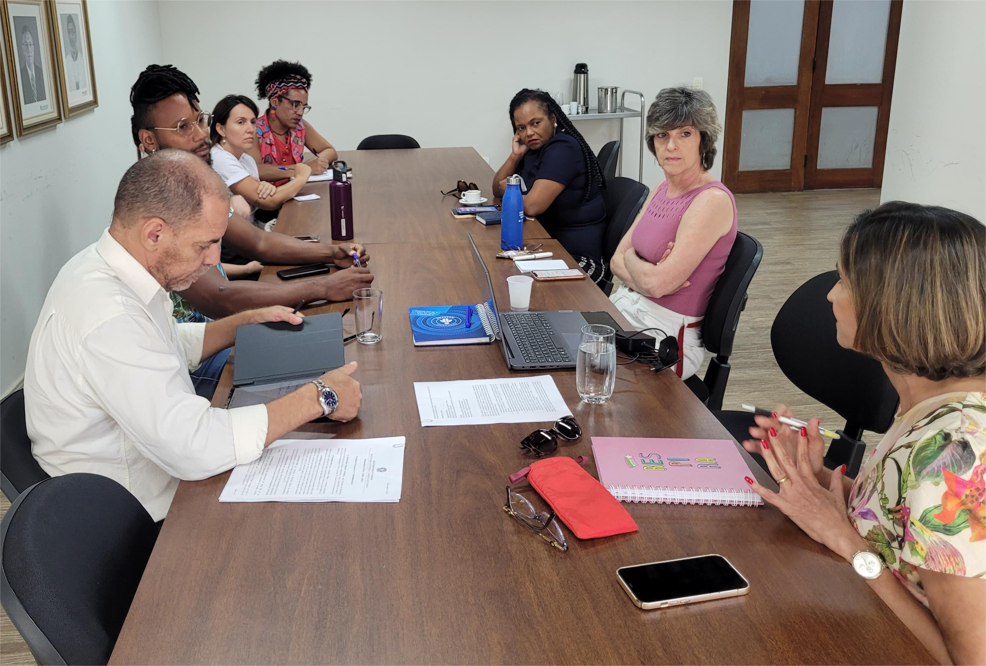 Foto da reunião, onde todos os participantes estão em uma sala de paredes brancas, sentados em torno de uma grande mesa de madeira 