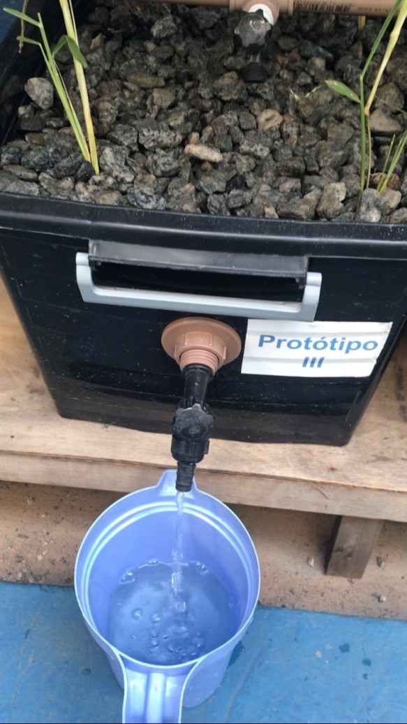 Foto de uma caixa de biocarvão com uma torneira de onde sai água limpa caindo em um recipiente azul