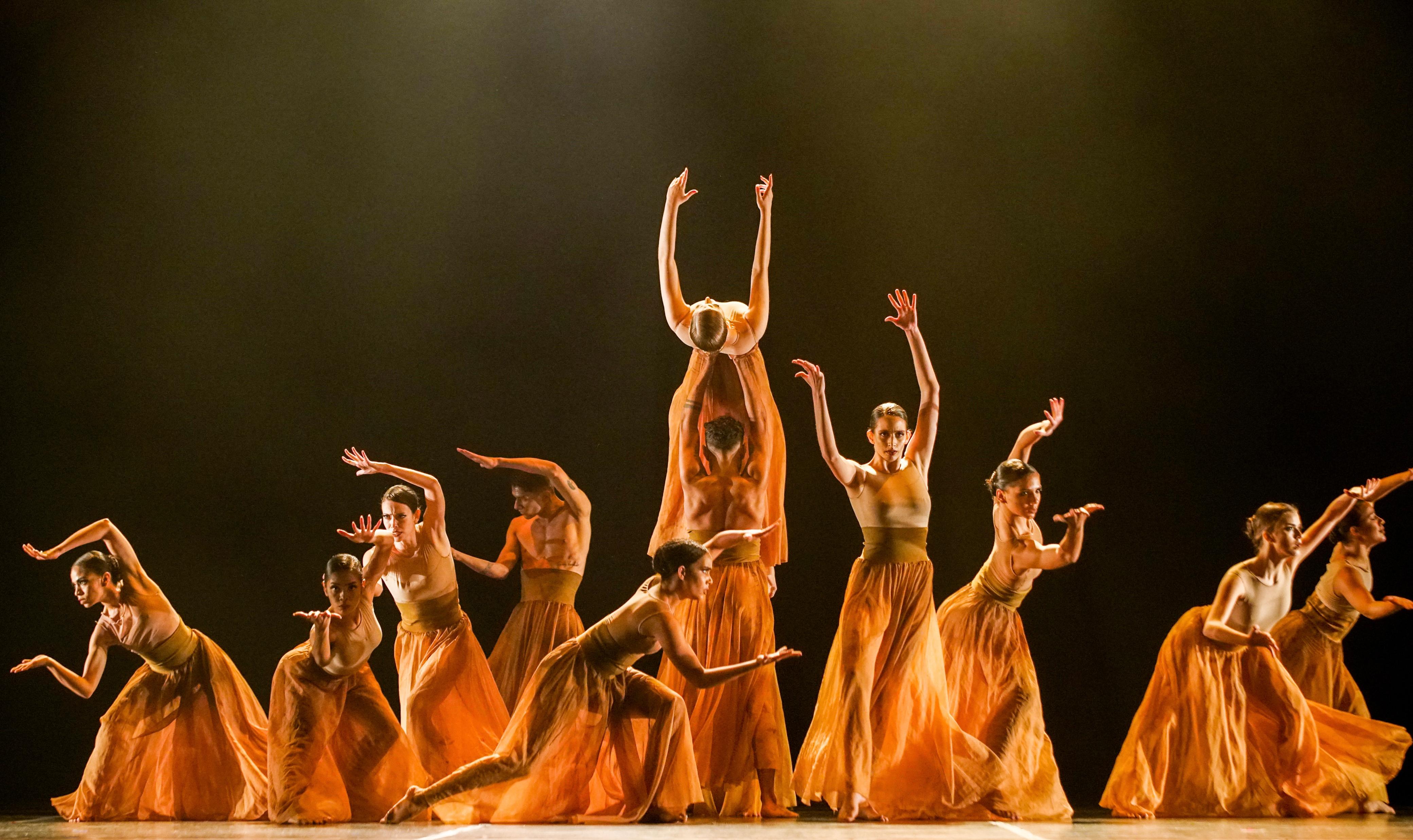 Foto de um grupo de bailarinos vestidos com roupas predominantemente na cor laranja se apresentando em um palco.