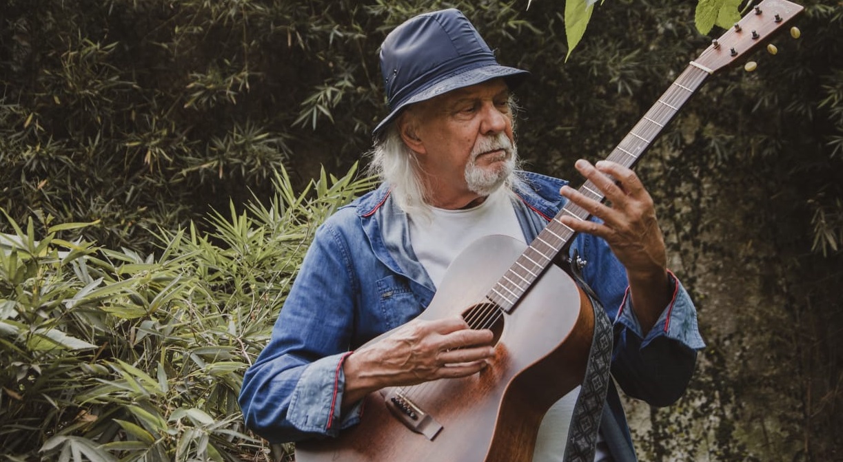 Foto do cantor Zé Geraldo tocando violão em uma área verde.