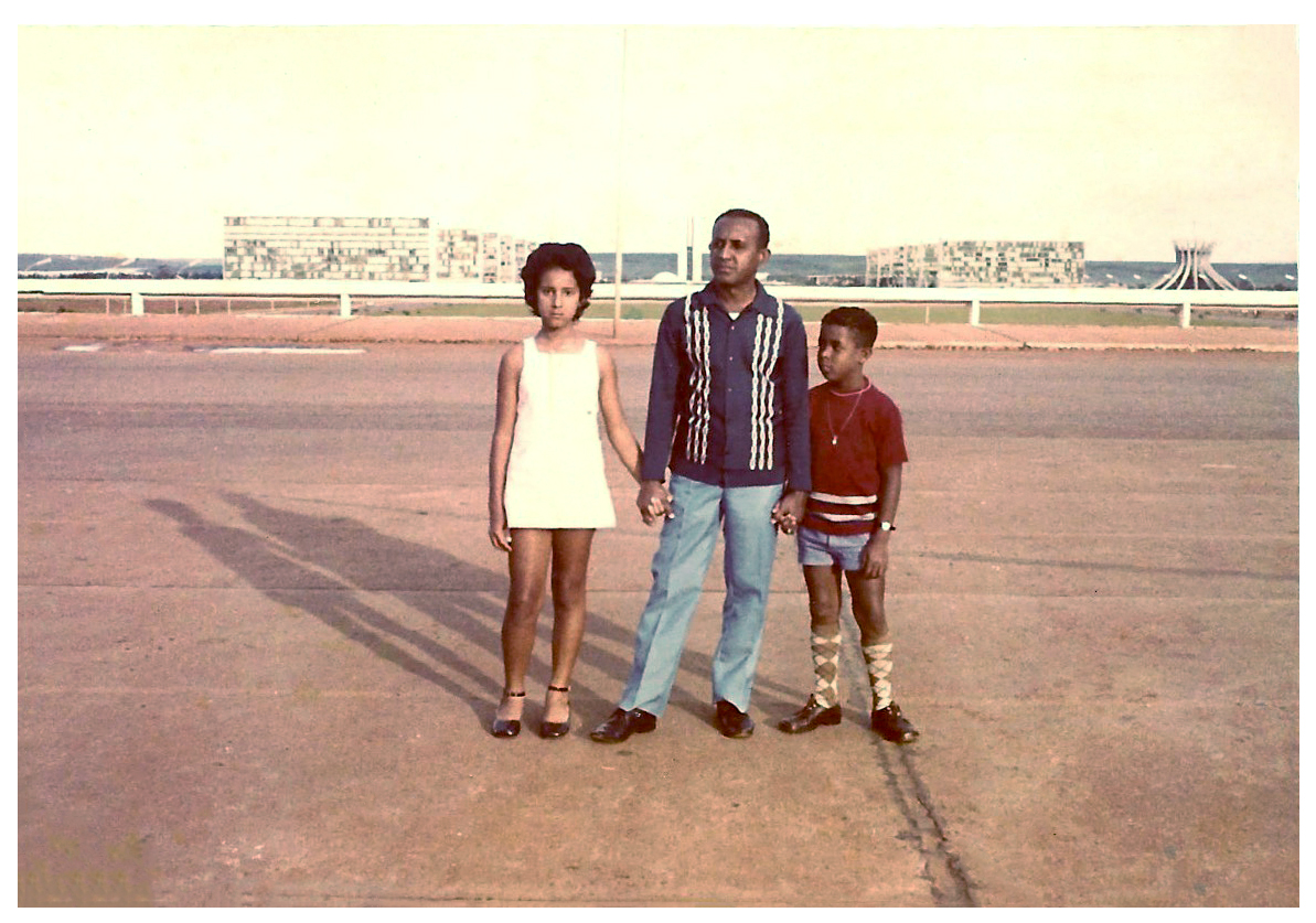 Imagem de uma cena do filme A Serena Onda que o Mar me Trouxe. Nela, em uma calçada, um homem segura nas mãos um menino e uma menina. Os três posam para uma foto, tendo ao fundo a imagem da Praça dos Três Poderes, em Brasília.
