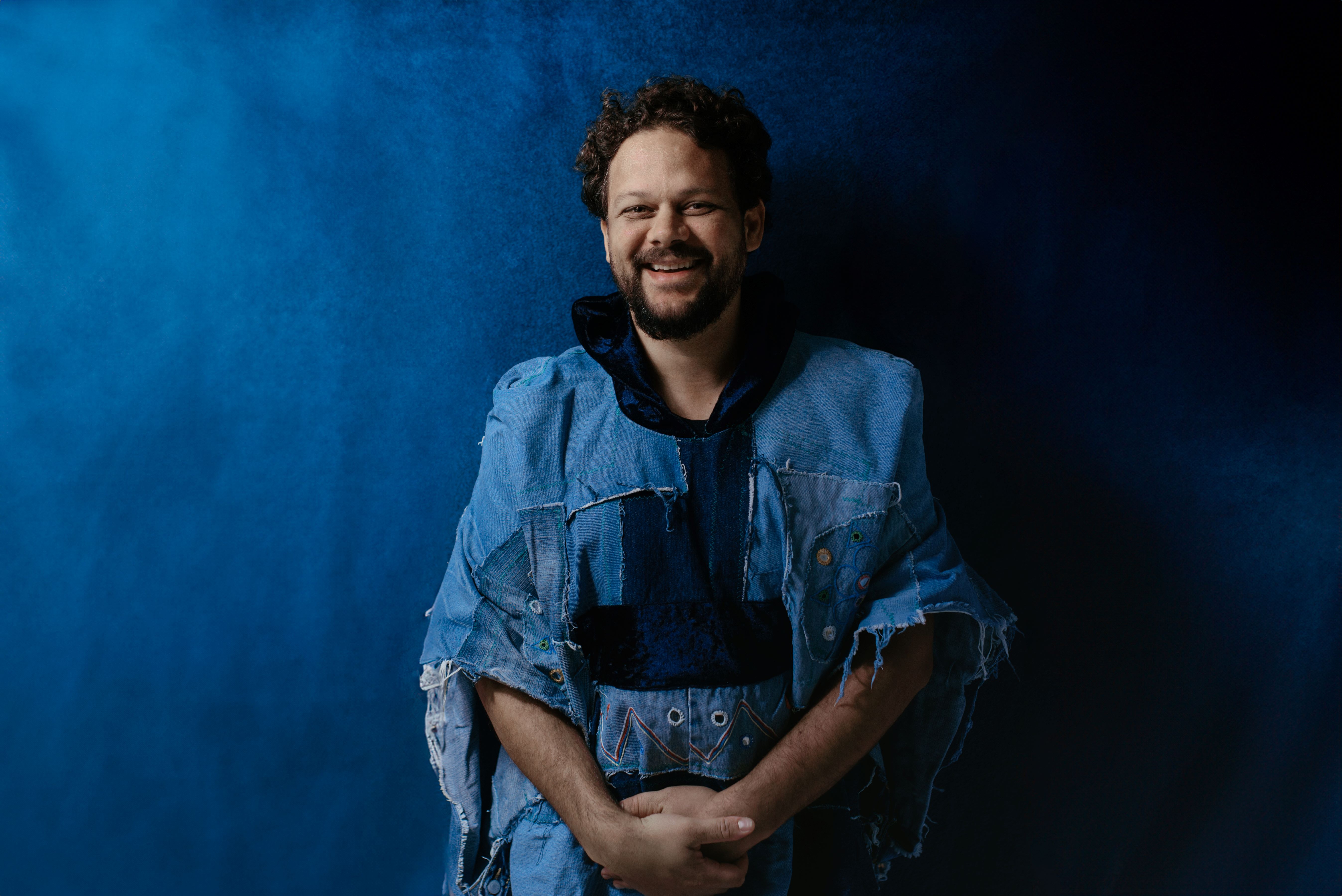 Foto do cantor em pé, sorrindo e posando em um cenário com fundo azul