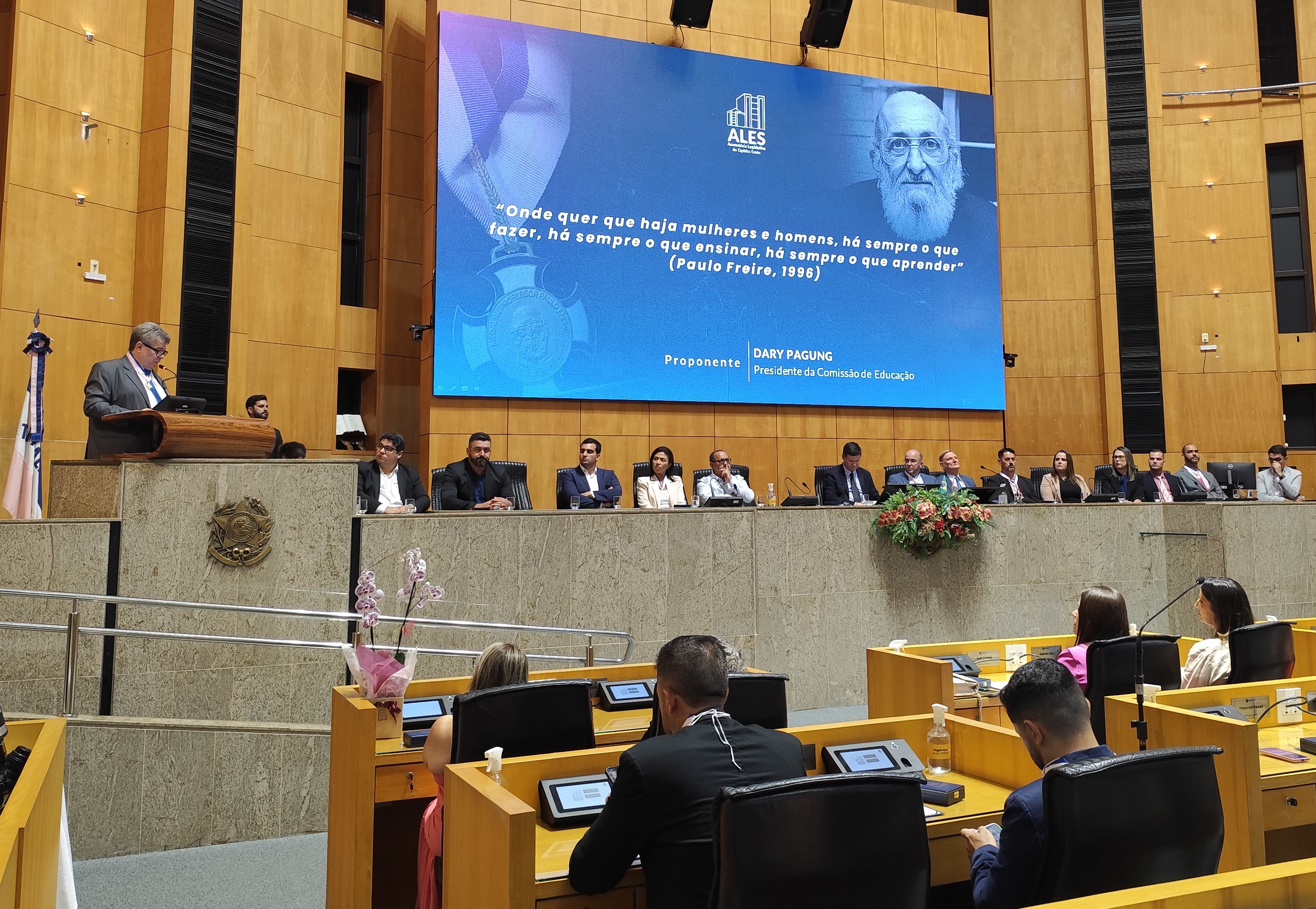 Foto do reitor no púlpito da Assembleia Legislativa do Espírito Santo. Ao fundo, a mesa principal com autoridades e, ao fundo, um telão no qual está projetada a figura de Paulo Freire.