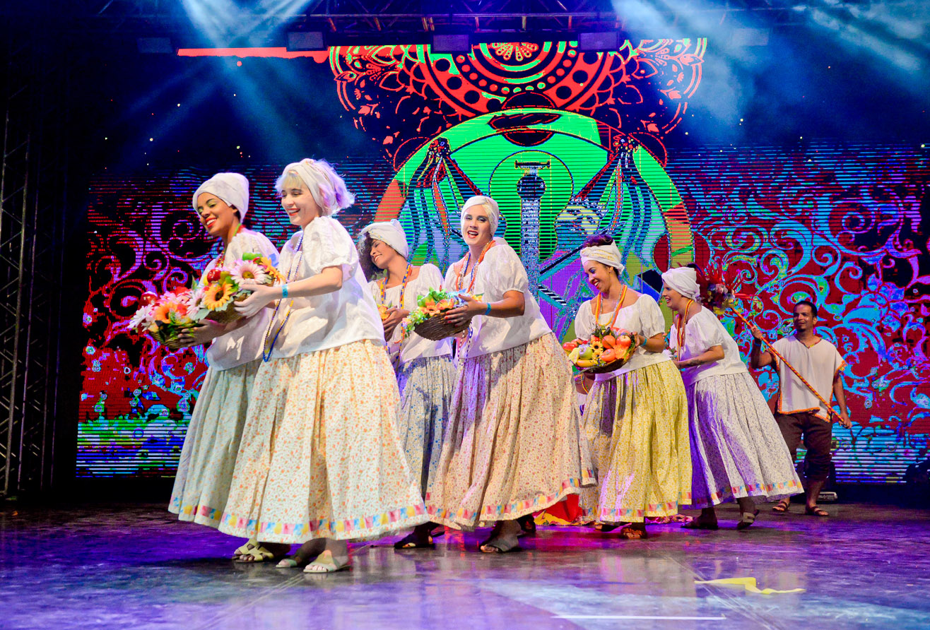 Foto de mulheres do Grupo Andora em uma apresentação no palco. Elas vestem blusas brancas, saias rodadas com estampa florida em tom amarelo claro. Nas mãos carregam flores como oferendas..