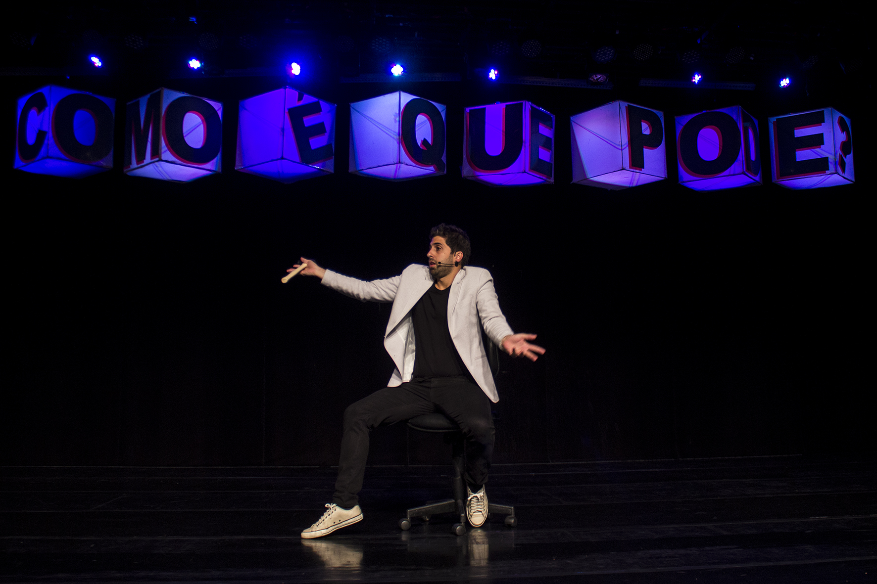 Foto do humorista no palco. Atrás dele, cubos iluminados de azul formam a frase "como é que pode?"