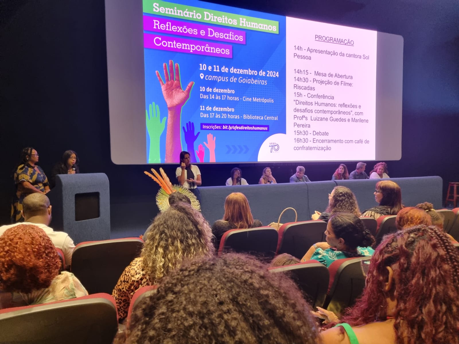 Foto da mesa abertura do evento, com o cartaz de divulgação projetado na tela do cinema