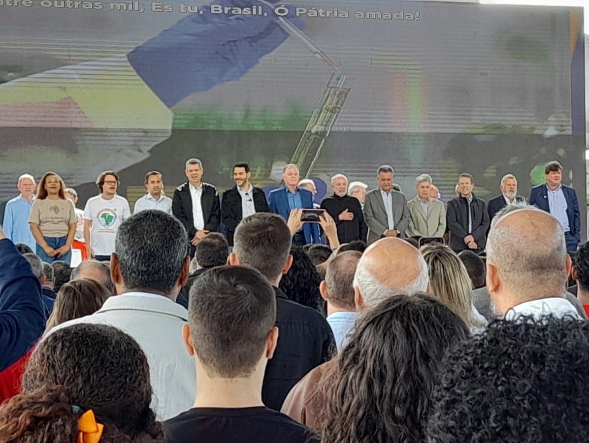 Foto da solenidade, com as pessoas em pé cantando o Hino Nacional