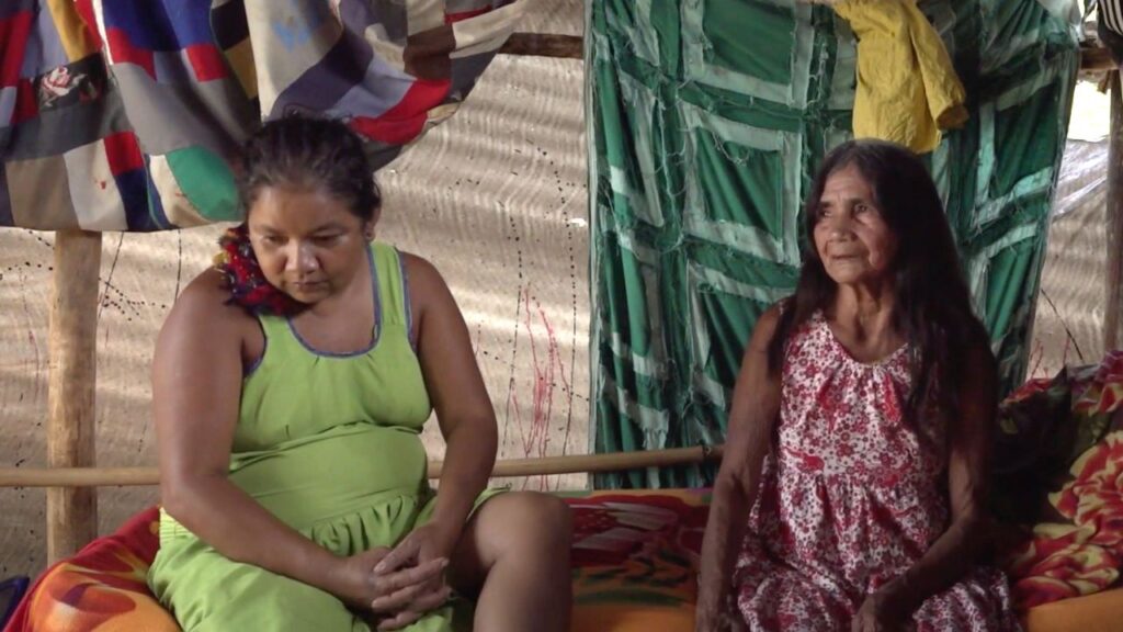 Cena de Yõg Ãtak: Meu Pai, Kaiowá na qual aparecem duas mulheres indígenas sentadas no interior do que parece ser uma oca