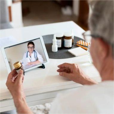 Foto de paciente homem de costas, sentado, sendo atendido por médica através de tablet, que está sobre a mesa