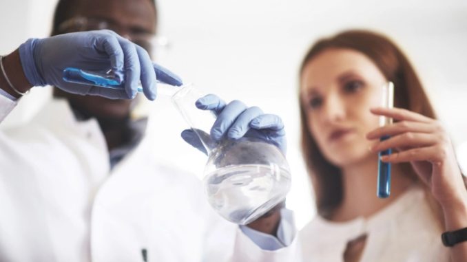 Foto de um homem negro e uma mulher branca, ambos usando roupas brancas e segurando materiais de laboratório