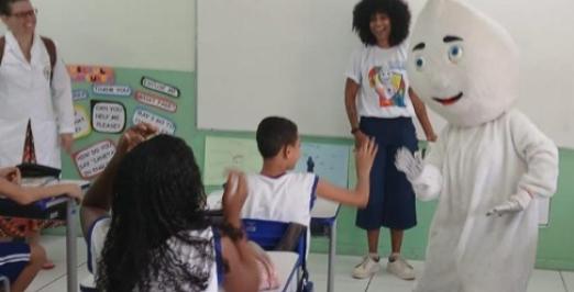 Foto de uma sala de aula onde uma pessoa vestida de Zé Gotinha cumprimenta um aluno