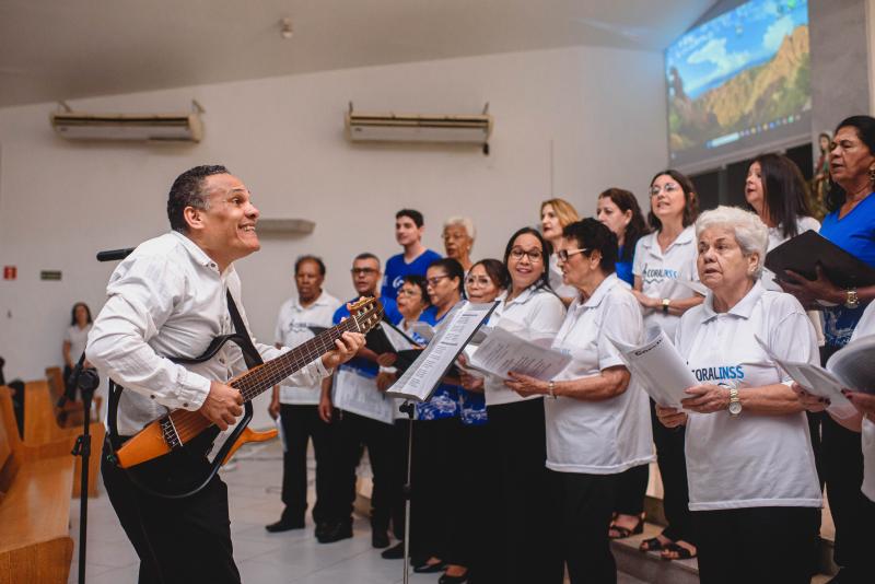 Foto do Coral do INSS, com os coralistas em uma apresentação. À frente do grupo, o maestro toca guitarra.