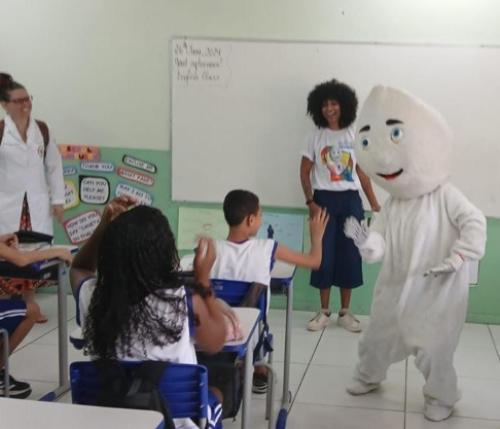 Foto de uma sala de aula, onde uma pessoa vestida de Zé Gotinha cumprimenta um aluno