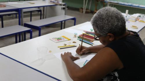 Foto de uma senhora fazendo o desenho de uma joia. Sobre a mesa, papel e lápis de cor 