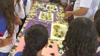 Crianças uniformizadas em volta de uma mesa olhando miniaturas de insetos, atentas à explicação de um monitor