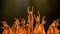 Foto de um grupo de bailarinos vestidos com roupas predominantemente na cor laranja se apresentando em um palco.