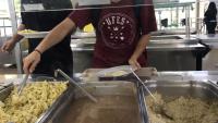 Foto de um estudante, vestindo uma camiseta da Ufes, se servindo diante do balcão do restaurante universitário. O rosto do estudante não aparece