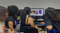 Foto de estudantes sentados em torno de um computador, olhando para a tela
