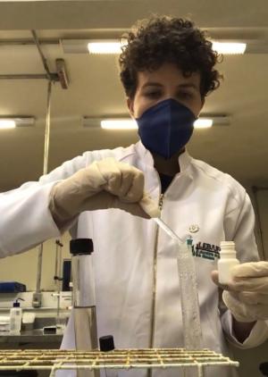 Pesquisadora com jaleco e máscara em um laboratório colocando alguma substância dentro de um tubo de ensaio