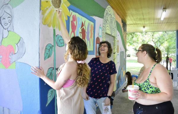 A foto mostra três participantes do projeto em frente ao painel. Uma delas pinta um girassol, enquanto as outras duas sorriem olhando o trabalho