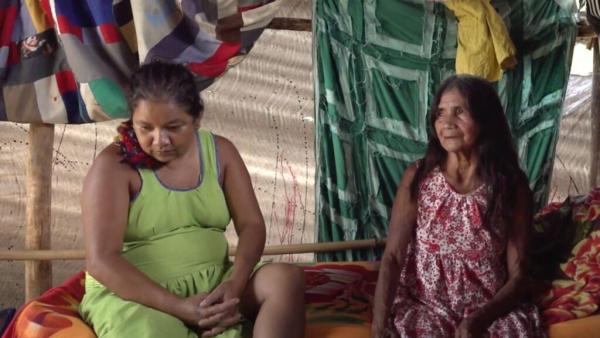 Cena de Yõg Ãtak: Meu Pai, Kaiowá na qual aparecem duas mulheres indígenas sentadas no interior do que parece ser uma oca