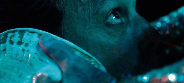 Cena de Peixe Abissal, na qual parte do rosto de um homem aparece emergindo de bolhas em um cenário azul