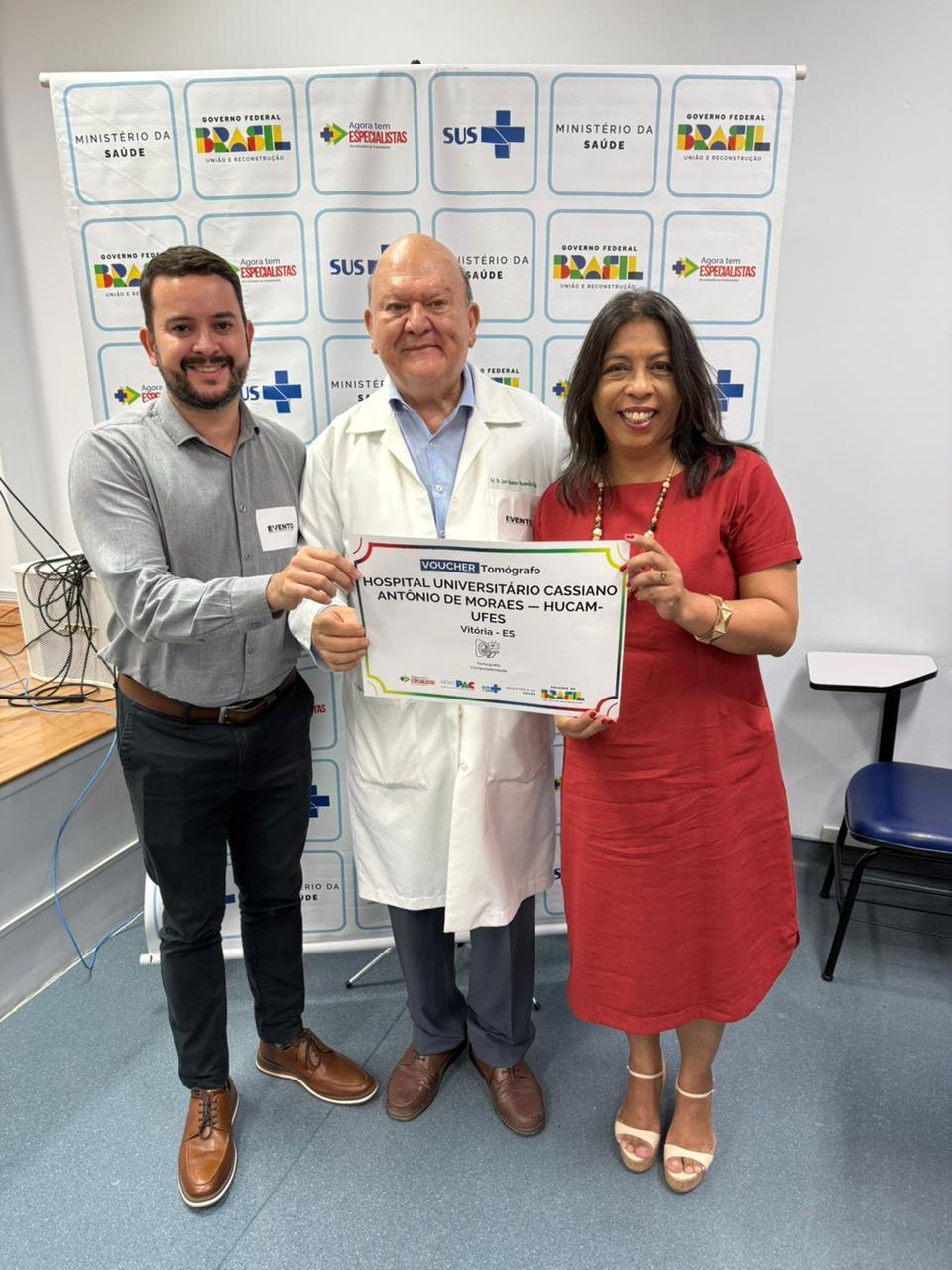 Foto do gerente administrativo do Hucam, César Dias, do superintendente do Hospital e da vice-reitora segurando um "voucher tomógrafo"