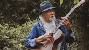 Foto do cantor Zé Geraldo tocando violão em uma área verde.