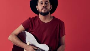 Foto do músico Rodrigo Suricato usando um chapéu preto e segurando um violão 