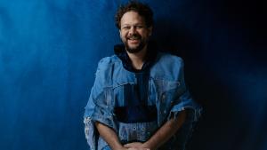 Foto do cantor em pé, sorrindo e posando em um cenário com fundo azul