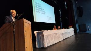 Foto do professor David Rodrigues, em primeiro plano, no púlpito do teatro. Ao fundo, a vice-reitora e a professora Denise Meyrelles sentadas à mesa principal