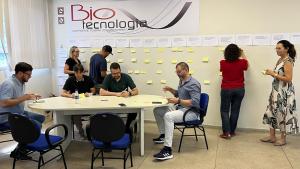 Grupo de participantes em uma mesa elaborando o planejamento enquanto outras pessoas fixam papeis na parede como parte da atividade