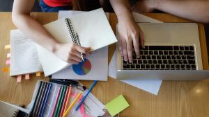 Imagem do alto de dois de estudantes diante de um notebook, caderno e canetas 