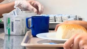 Foto de mãos segurando uma bandeja onde há um prato com um pão e uma xícara. Na imagem aparecem ainda duas mãos colocando um sachê de açúcar sobre a bandeja