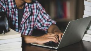Foto de um jovem digitando em um notebook sobre uma mesa com livros