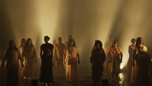 Foto dos cantores em pé, vestindo roupas em tons terrosos, se apresentando em um palco sob luzes amarelas