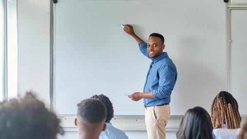 Professor em uma sala de aula escrevendo no quadro branco, de frente para a turma de jovens estudantes