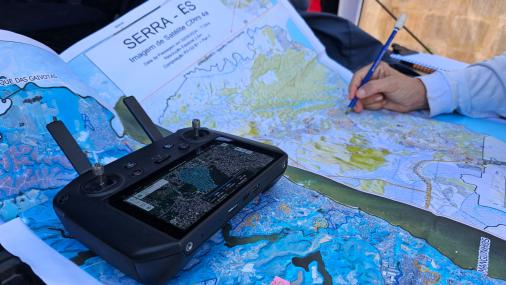 Foto de um mapa aberto do município da Serra com um controle remoto em cima e a mão de uma pessoa fazendo uma anotação a lápis sobre o mapa