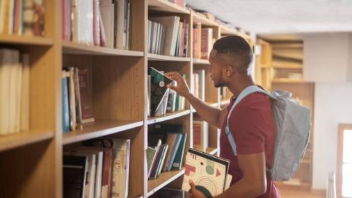 Foto de um estudante negro pegando um livro na estante de uma biblioteca